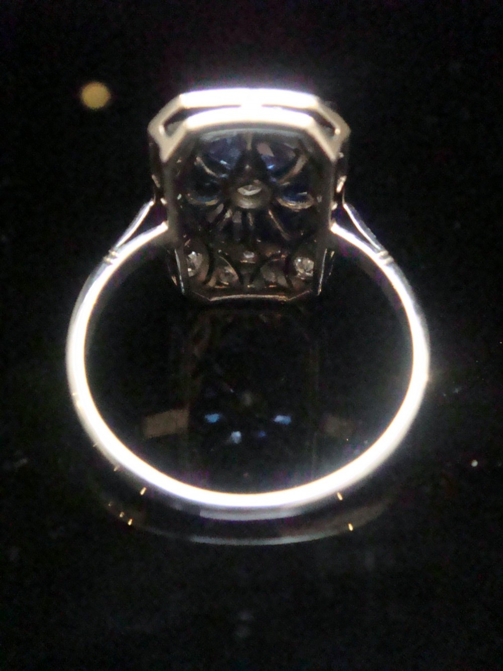 An Art Deco rectangular white gold ring featuring a central round diamond surrounded by calibre-cut blue sapphires and framed by smaller diamonds with detailed millegrain edging. View VIII