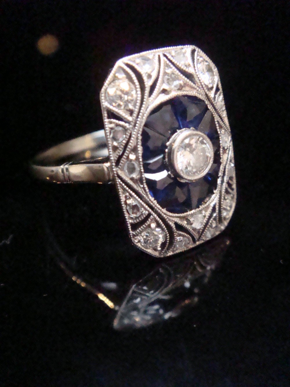 An Art Deco rectangular white gold ring featuring a central round diamond surrounded by calibre-cut blue sapphires and framed by smaller diamonds with detailed millegrain edging. View IX