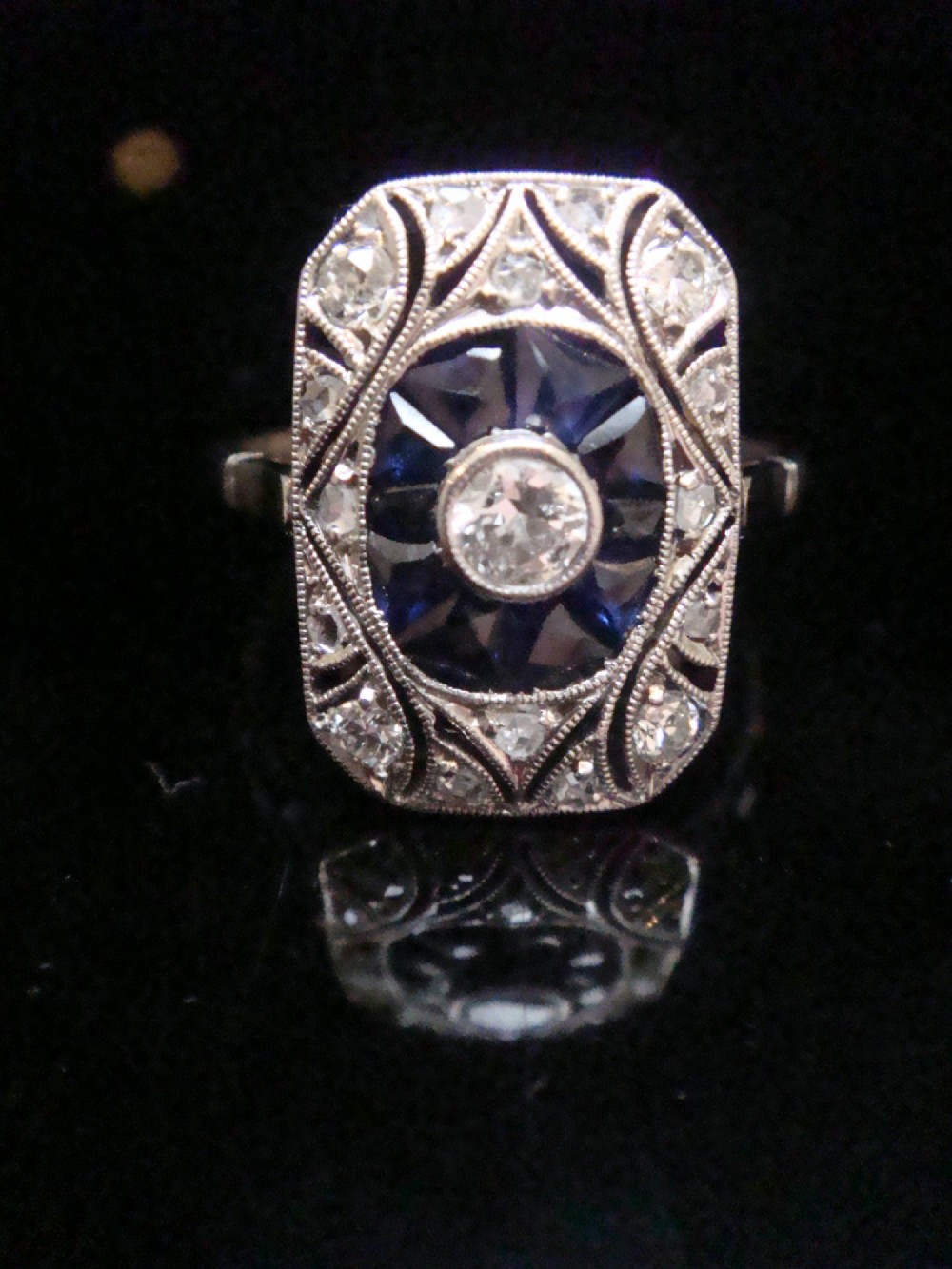 An Art Deco rectangular white gold ring featuring a central round diamond surrounded by calibre-cut blue sapphires and framed by smaller diamonds with detailed millegrain edging.