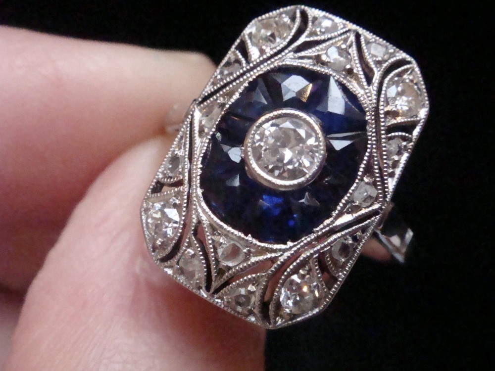 An Art Deco rectangular white gold ring featuring a central round diamond surrounded by calibre-cut blue sapphires and framed by smaller diamonds with detailed millegrain edging. View III