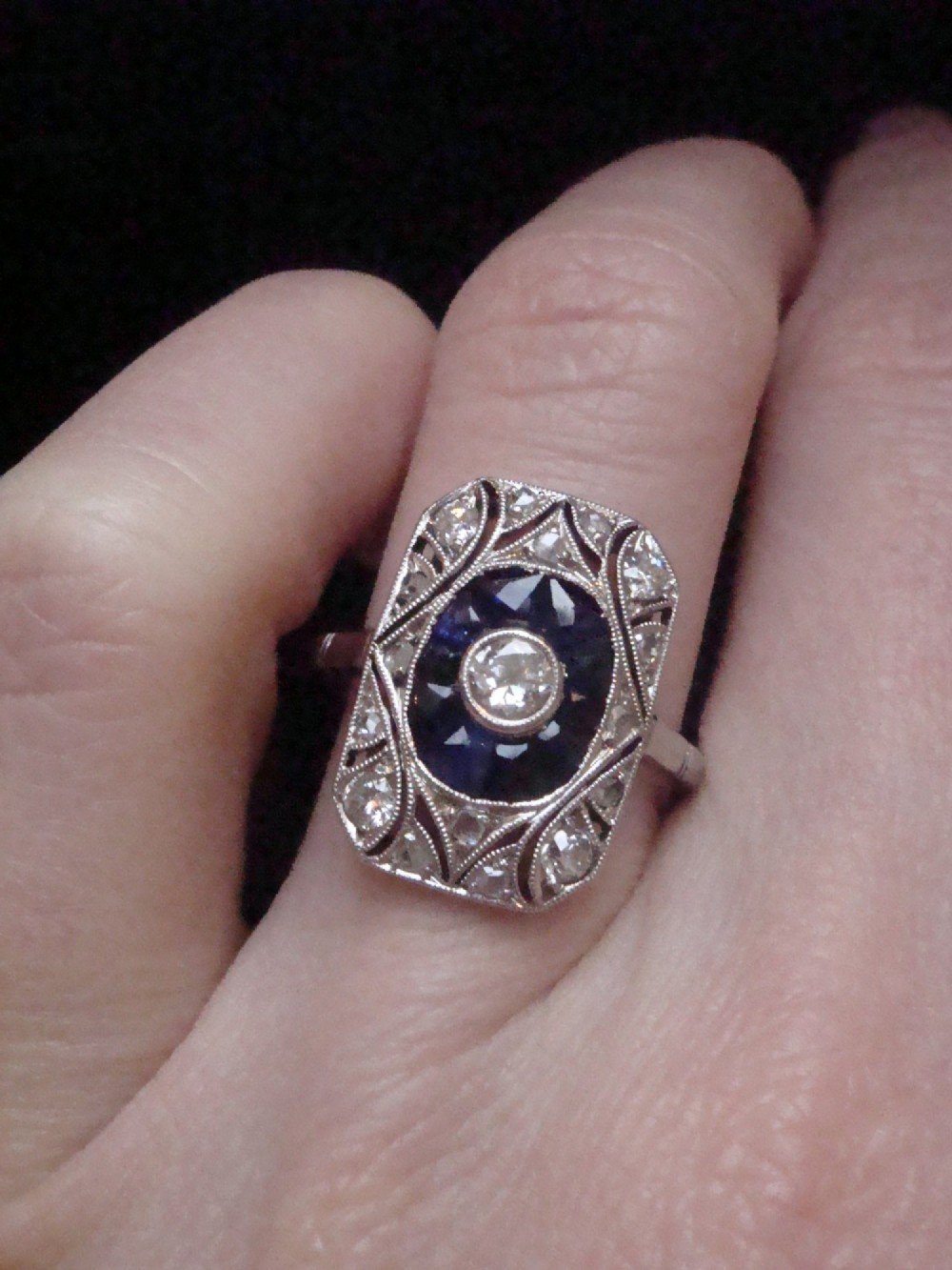 An Art Deco rectangular white gold ring featuring a central round diamond surrounded by calibre-cut blue sapphires and framed by smaller diamonds with detailed millegrain edging. View V