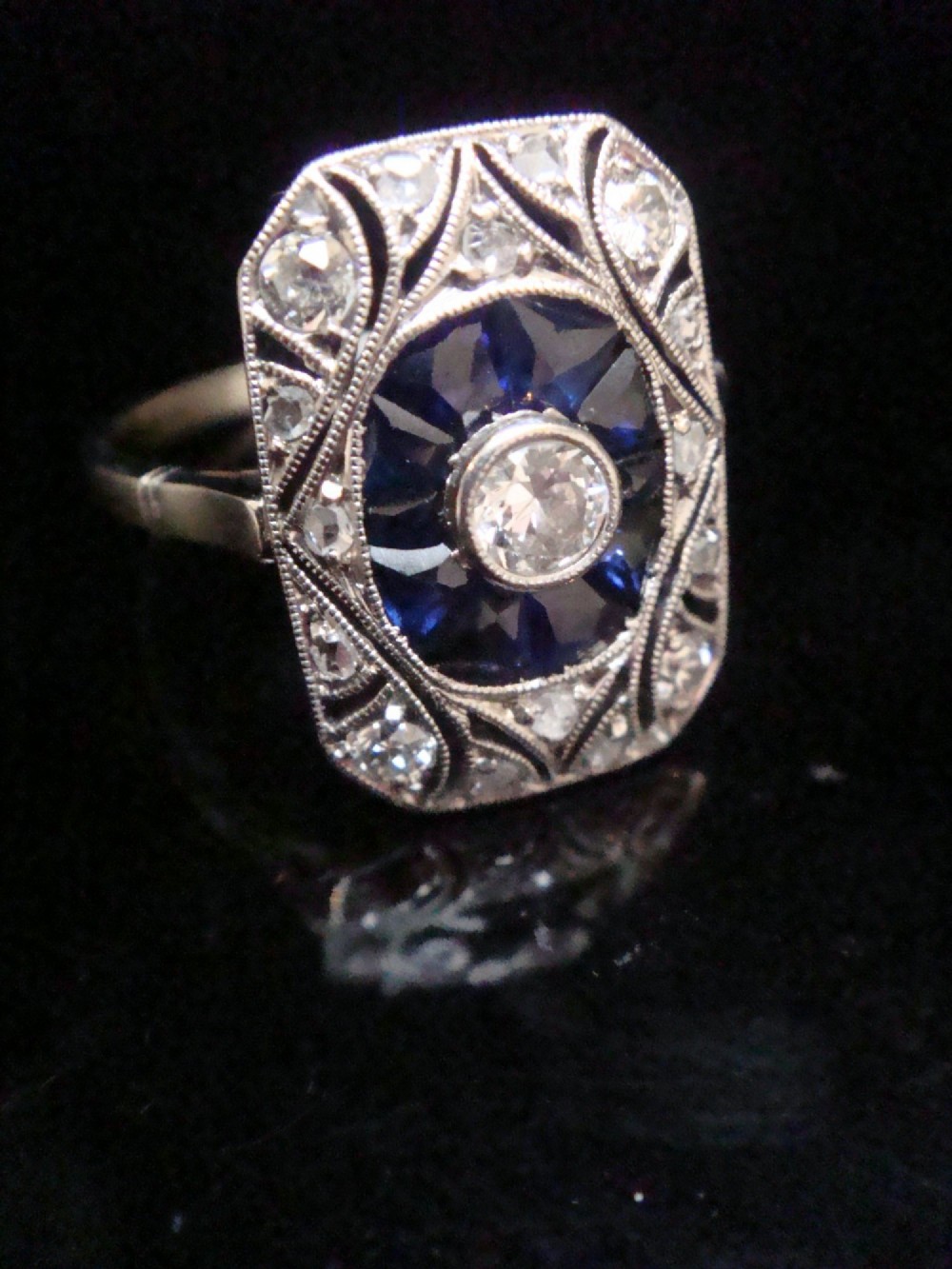An Art Deco rectangular white gold ring featuring a central round diamond surrounded by calibre-cut blue sapphires and framed by smaller diamonds with detailed millegrain edging. View VII