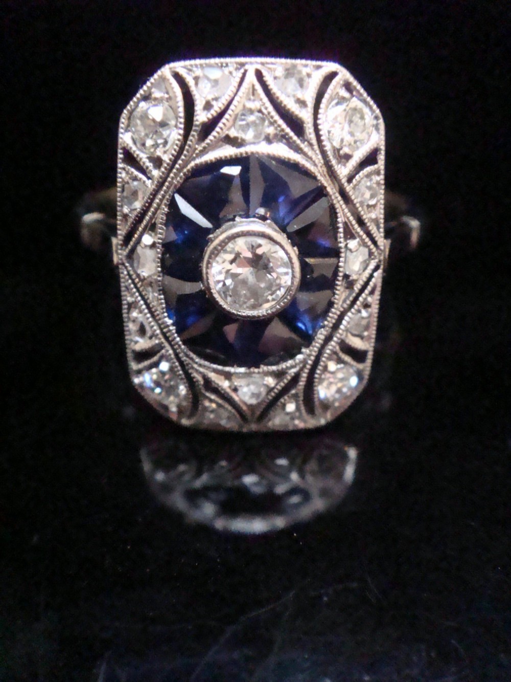 An Art Deco rectangular white gold ring featuring a central round diamond surrounded by calibre-cut blue sapphires and framed by smaller diamonds with detailed millegrain edging. View VI