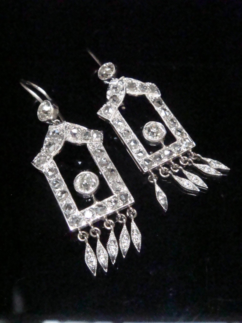A pair of platinum Art Deco drop earrings featuring openwork geometric frames set with old-cut diamonds and dangling diamond leaf fringes.