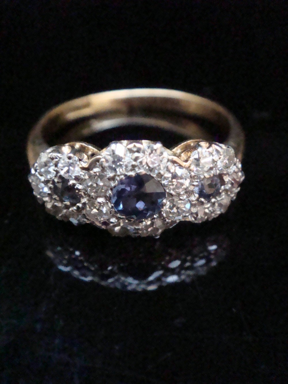 An Edwardian 18ct yellow gold ring featuring three cluster mounts set with dark blue sapphires and surrounded by small diamonds.