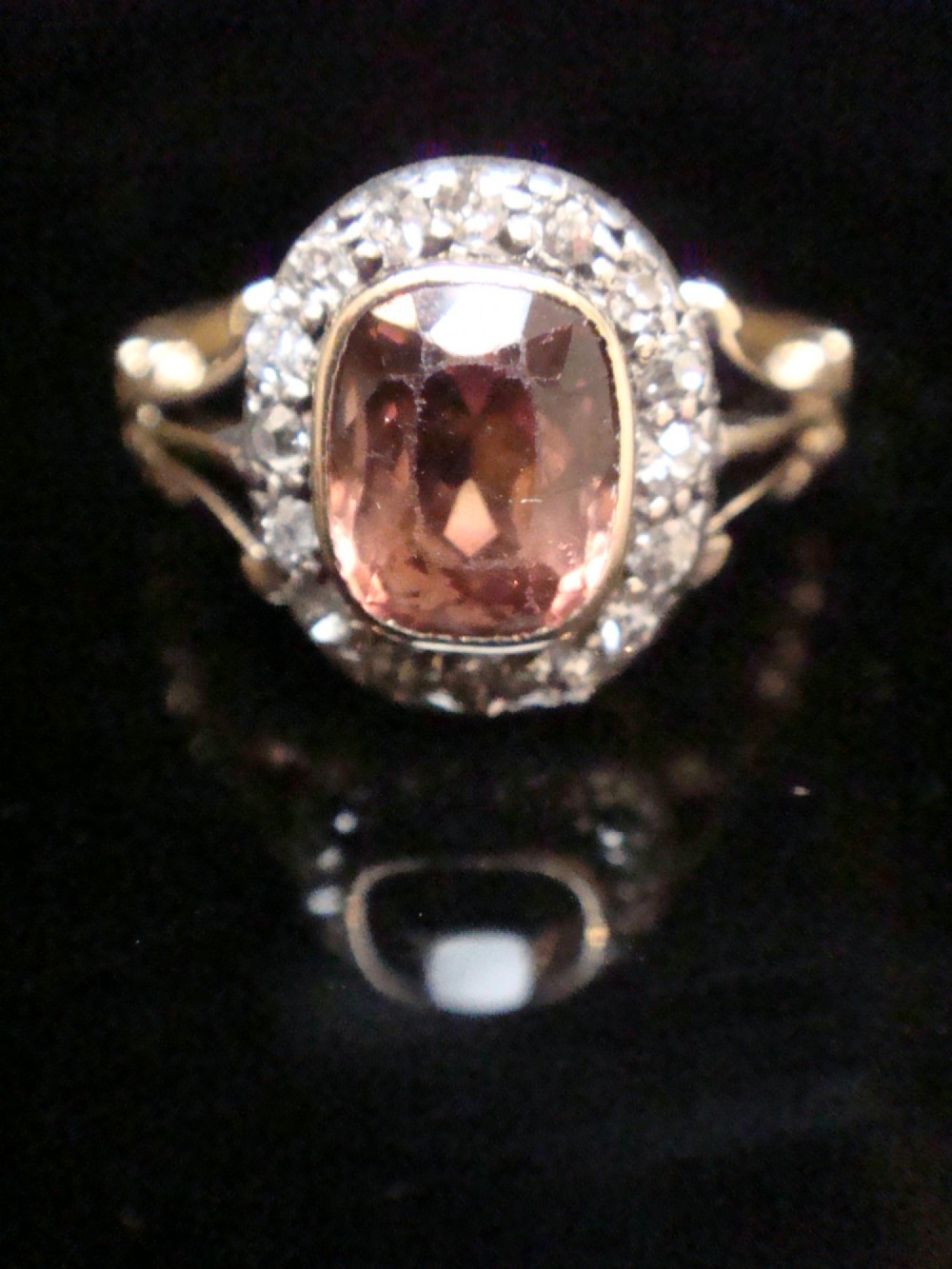 An Edwardian 18ct gold ring with a central cushion-cut peach tourmaline surrounded by a halo of old-cut diamonds and bifurcated shoulders. View V