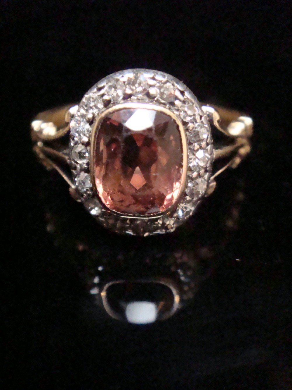 An Edwardian 18ct gold ring with a central cushion-cut peach tourmaline surrounded by a halo of old-cut diamonds and bifurcated shoulders. View VI