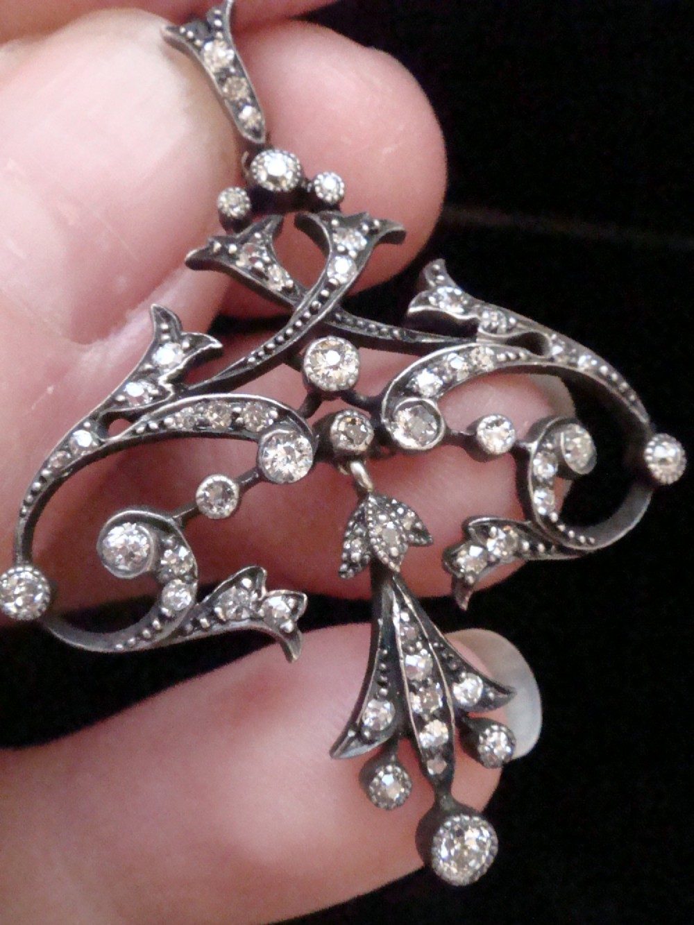 An openwork silver Edwardian lavaliere pendant set with round diamonds in a scrollwork design with a dangling central diamond drop. View IV