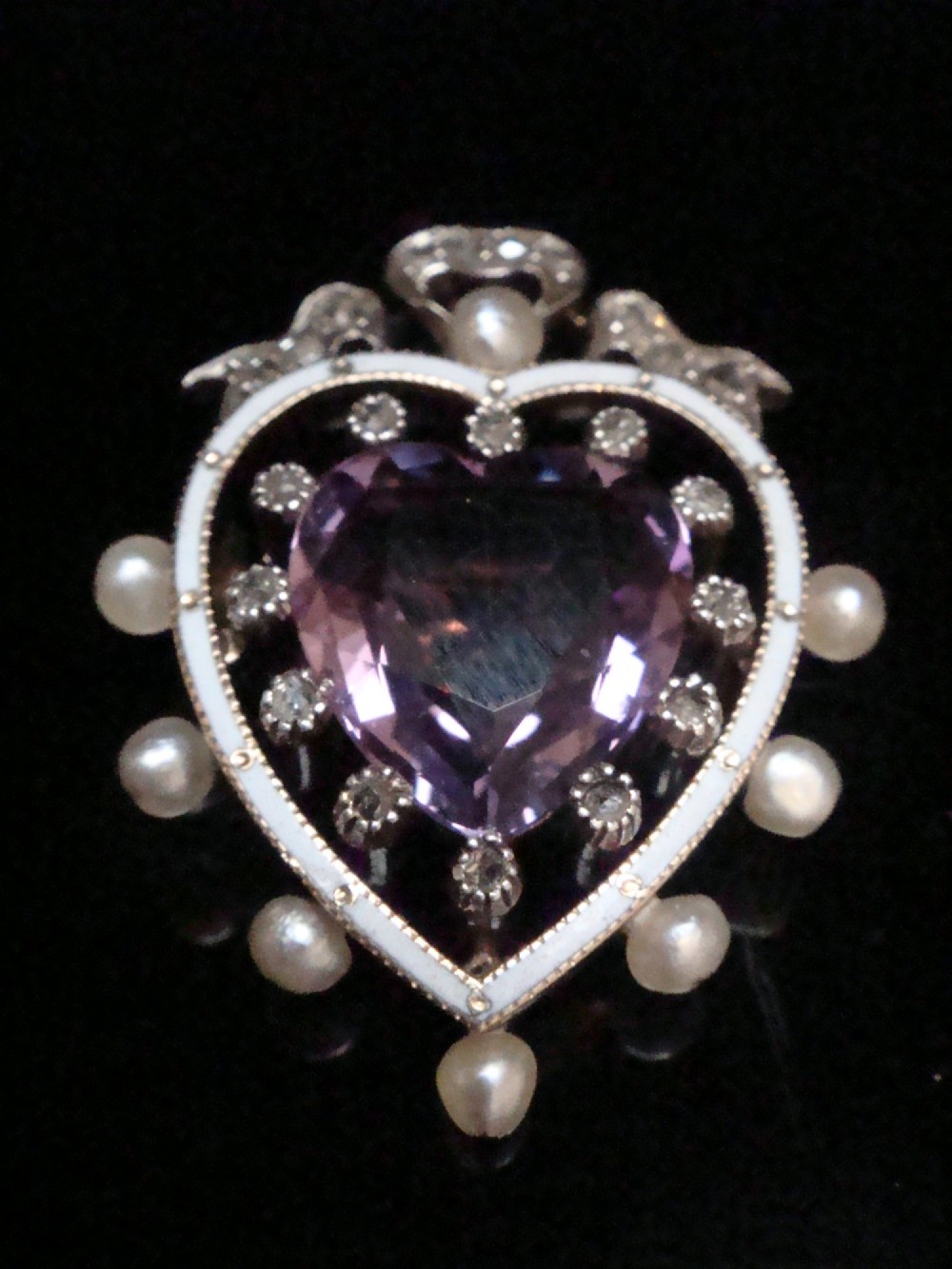 An Edwardian heart-shaped gold pendant featuring a central purple gemstone framed by diamonds, white guilloche enamel, and small natural pearls.