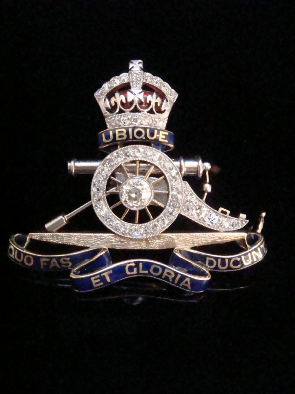 An Edwardian platinum and gold Royal Artillery badge brooch featuring blue guilloche enamel mottos and diamond settings.