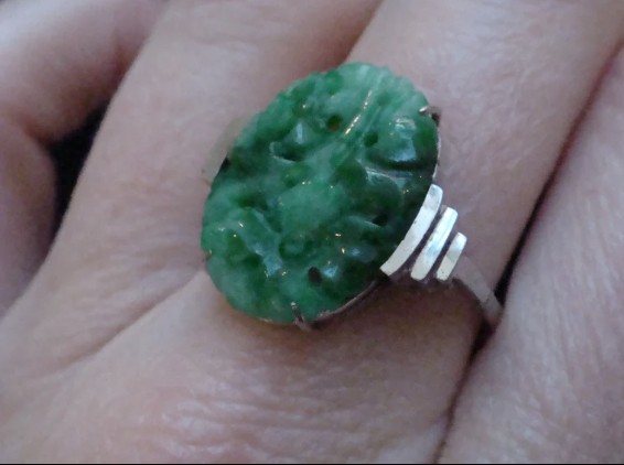 An Art Deco platinum ring featuring a carved oval imperial green jadeite gemstone set with stepped geometric shoulders. View IV