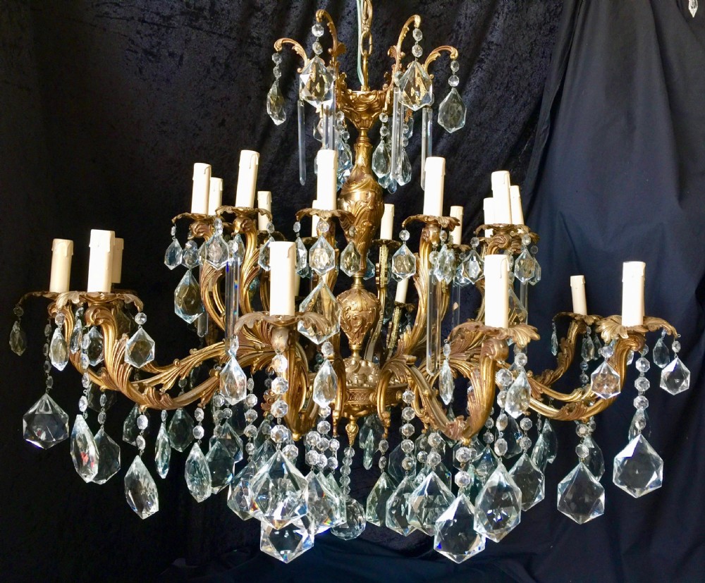 A large two-tiered brass chandelier featuring ornate scrolling arms, candle-effect sleeves, and clear faceted crystal droplets.