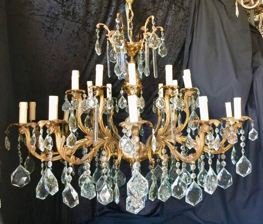 A large two-tiered brass chandelier featuring ornate scrolling arms, candle-effect sleeves, and clear faceted crystal droplets. View II