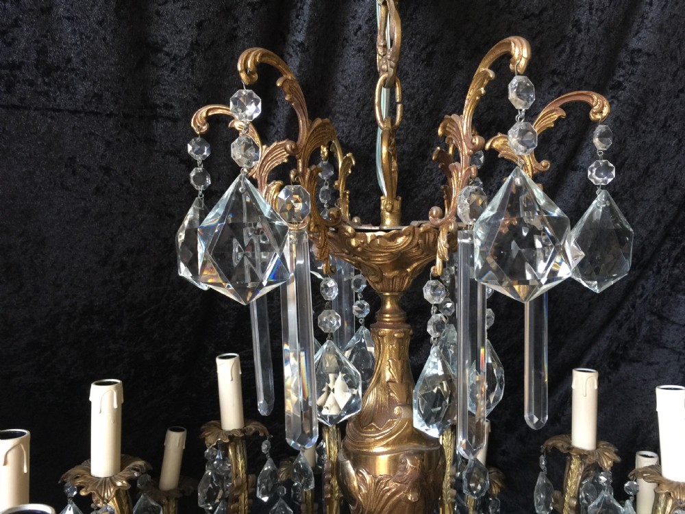 A large two-tiered brass chandelier featuring ornate scrolling arms, candle-effect sleeves, and clear faceted crystal droplets. View III