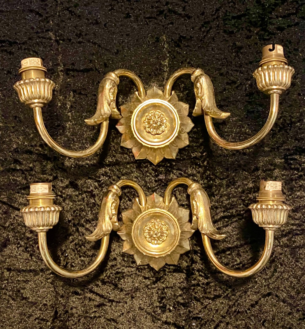 A pair of cast brass two-light wall sconces featuring sunburst backplates and curved acanthus leaf arms.