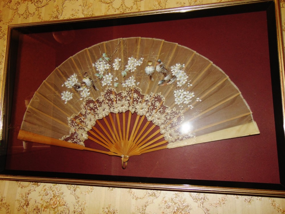 An Edwardian hand fan featuring painted birds and white blossoms on cream silk with lace trim, displayed within a framed Shadowbox frame.