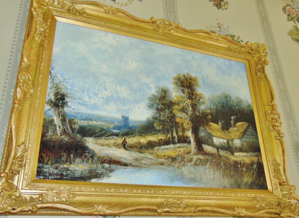 A late 19th-century oil painting on canvas depicting a rural landscape with thatched cottages and a church spire, housed in a heavily carved giltwood frame.