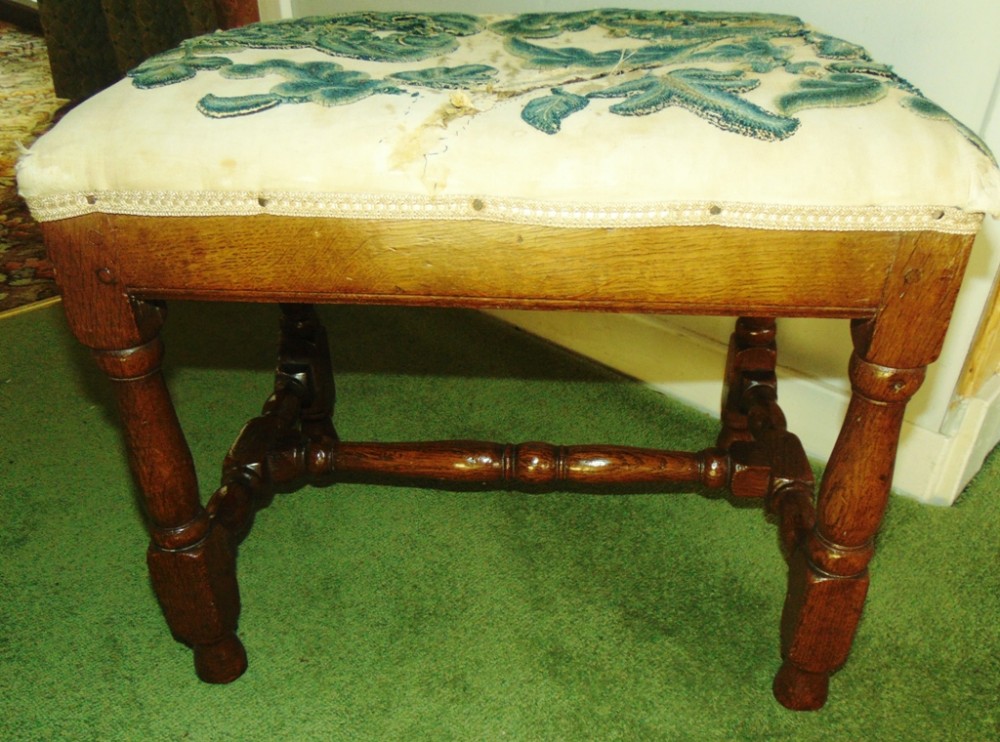 An 18th-century French oak stool with turned legs, a lower H-stretcher, and an off-white fabric seat featuring teal wool crewel work embroidery.