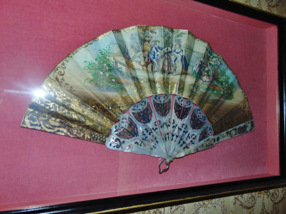 An antique Edwardian hand fan featuring painted pastoral figures and pierced mother-of-pearl sticks, displayed within a glazed frame.