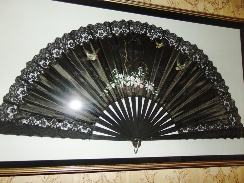 An antique Edwardian black silk folding fan featuring lace edging and hand-painted bird and floral motifs, displayed in a frame.