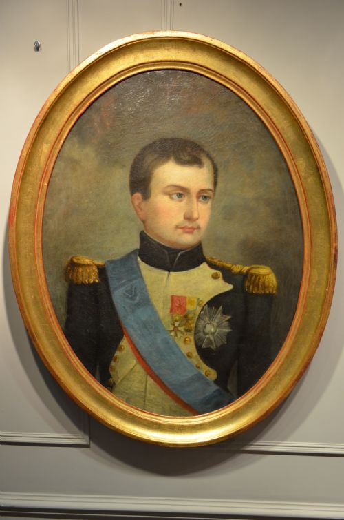 fig 1. An oval oil painting depicting Napoleon Bonaparte in a dark military uniform with gold epaulettes and a light blue sash, set within a gilt wood frame.