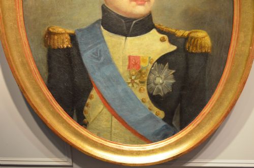fig 3. An oval oil painting depicting Napoleon Bonaparte in a dark military uniform with gold epaulettes and a light blue sash, set within a gilt wood frame.
