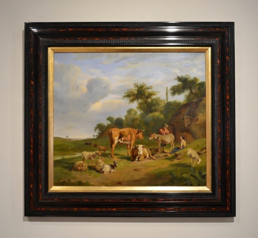 An antique framed Dutch pastoral oil painting depicting cattle, sheep, goats, a donkey, and figures in an open landscape under a cloud-filled sky.
