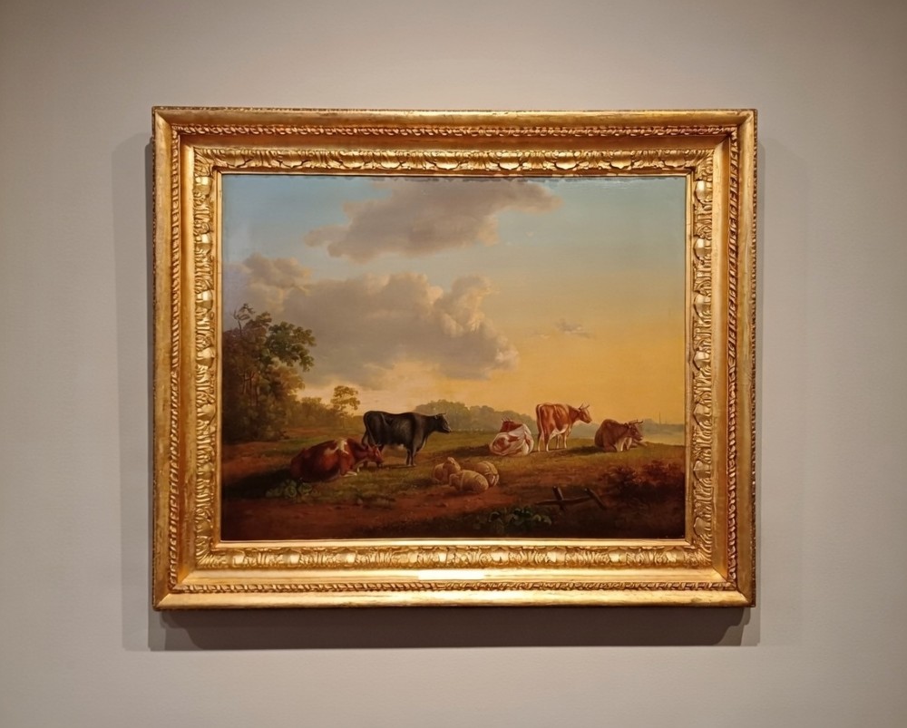 A 19th-century oil painting depicting cattle and sheep resting in an open pastoral landscape beneath a cloud-filled sky, enclosed in an ornate gilded frame.