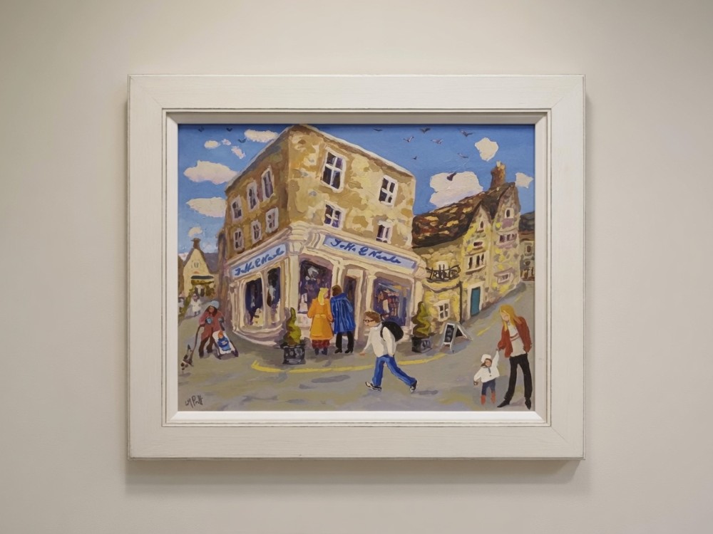 A framed original oil painting depicting the Jaffe and Neale shopfront on a Cotswold street corner with figures walking by, set in an off-white wooden frame.