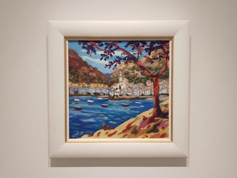 A framed original oil painting depicting a Mediterranean coastal village with blue water and a foreground tree with magenta leaves.