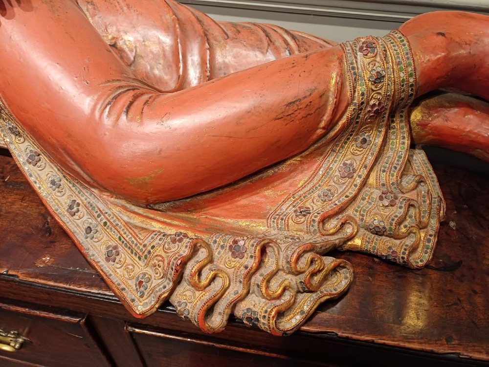 A 19th-century Burmese carved wooden sculpture of a reclining Buddha featuring red lacquered finish and detailed gilded robes. View VI