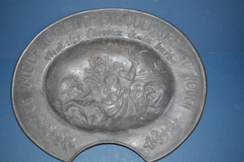 A 19th-century French pewter oval barber bowl with a crescent neck cut-out, embossed rim lettering, and a central relief scene.