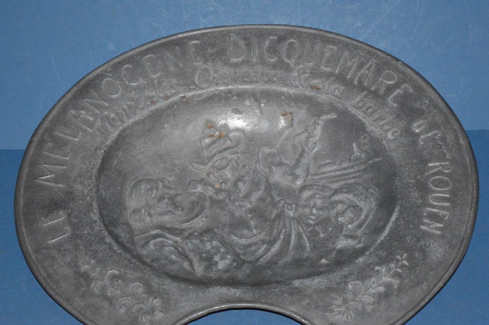 A 19th-century French pewter oval barber bowl with a crescent neck cut-out, embossed rim lettering, and a central relief scene. View II