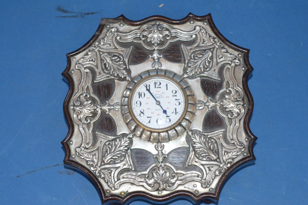 An Art Nouveau shaped wooden mantel clock clad in ornate chased silver relief with foliage motifs surrounding a central white enamel dial.