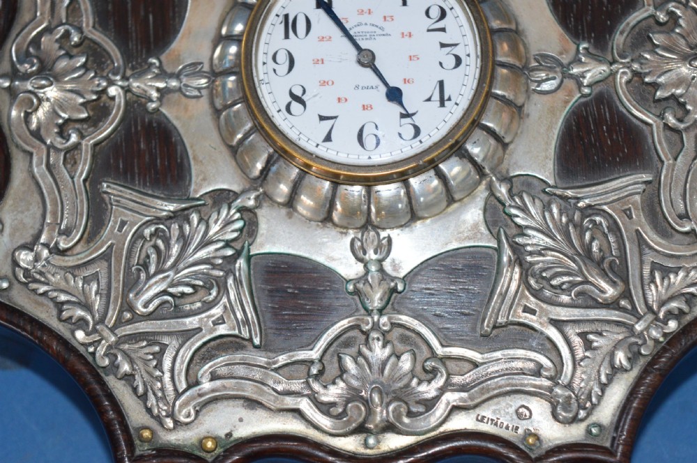 An Art Nouveau shaped wooden mantel clock clad in ornate chased silver relief with foliage motifs surrounding a central white enamel dial. View II