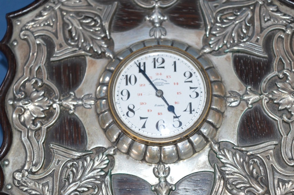 An Art Nouveau shaped wooden mantel clock clad in ornate chased silver relief with foliage motifs surrounding a central white enamel dial. View III