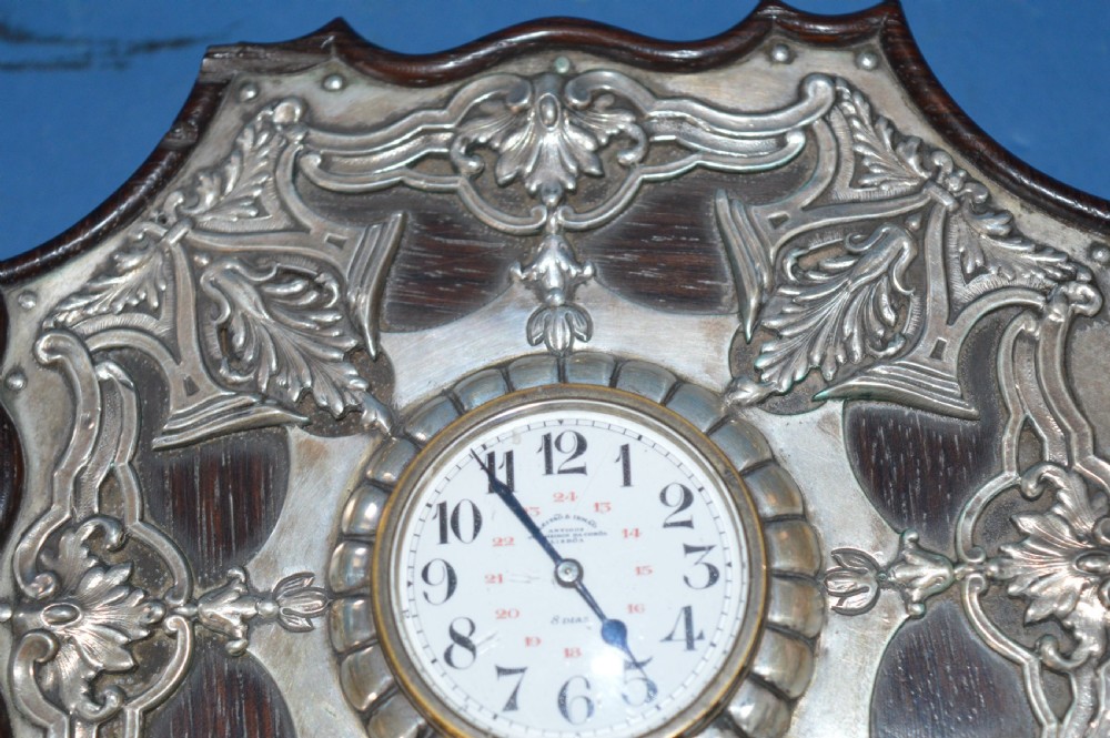 An Art Nouveau shaped wooden mantel clock clad in ornate chased silver relief with foliage motifs surrounding a central white enamel dial. View IV