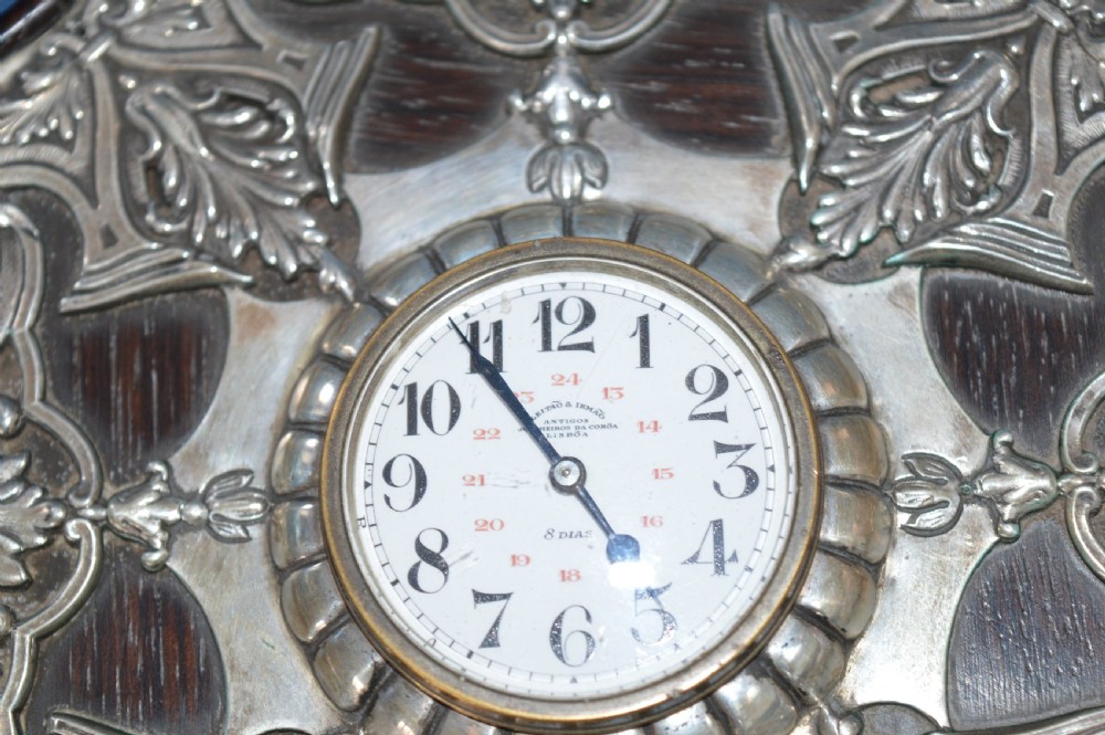 An Art Nouveau shaped wooden mantel clock clad in ornate chased silver relief with foliage motifs surrounding a central white enamel dial. View V
