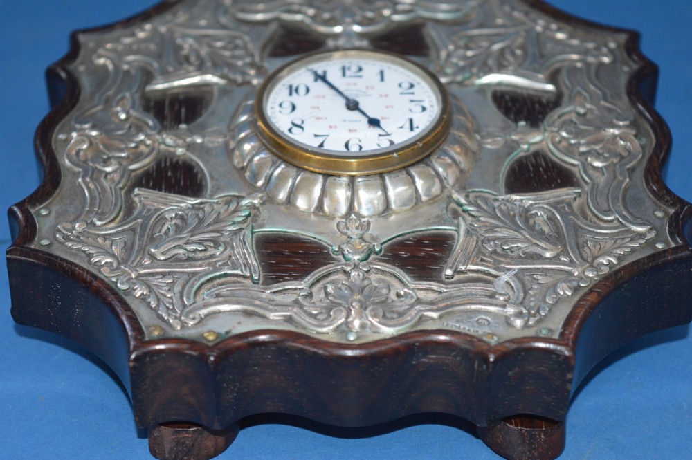 An Art Nouveau shaped wooden mantel clock clad in ornate chased silver relief with foliage motifs surrounding a central white enamel dial. View IX