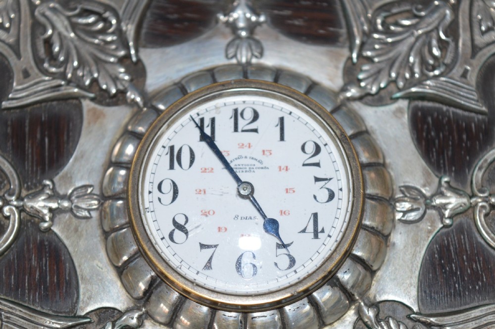 An Art Nouveau shaped wooden mantel clock clad in ornate chased silver relief with foliage motifs surrounding a central white enamel dial. View X