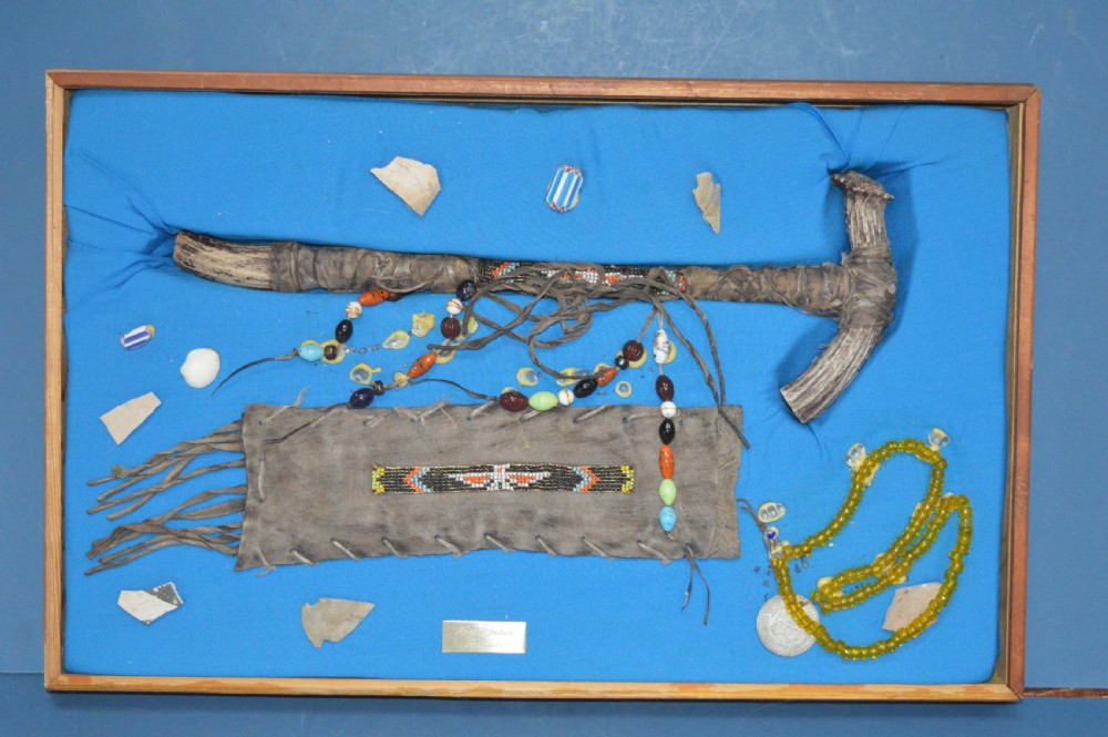 A framed display case lined with blue cloth containing Native American artefacts, including a beaded leather pouch, trade beads, arrowheads, and a hafted tool.