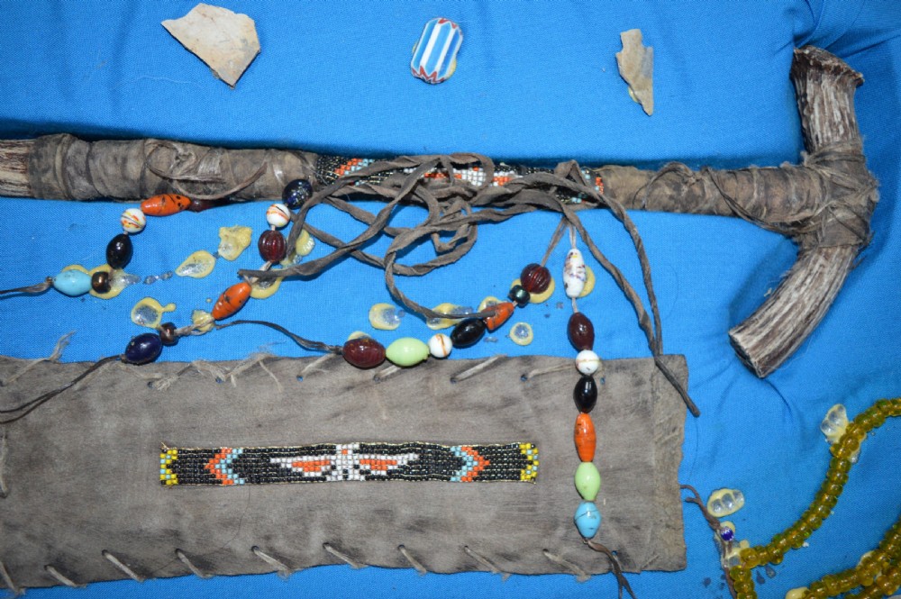 A framed display case lined with blue cloth containing Native American artefacts, including a beaded leather pouch, trade beads, arrowheads, and a hafted tool. View II