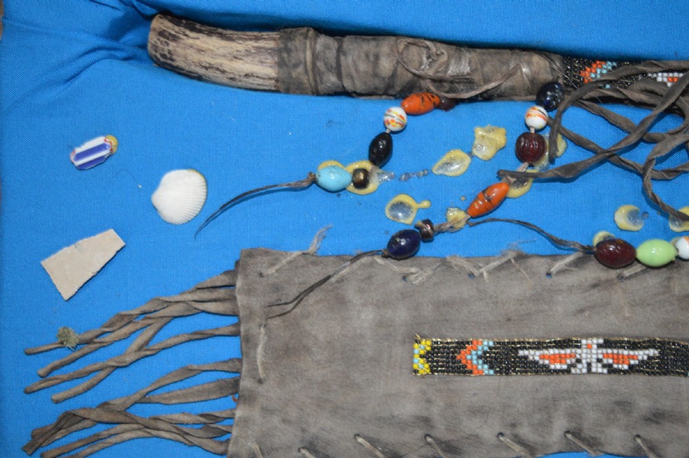 A framed display case lined with blue cloth containing Native American artefacts, including a beaded leather pouch, trade beads, arrowheads, and a hafted tool. View III