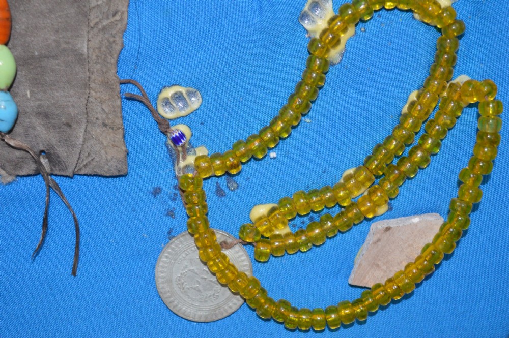 A framed display case lined with blue cloth containing Native American artefacts, including a beaded leather pouch, trade beads, arrowheads, and a hafted tool. View V