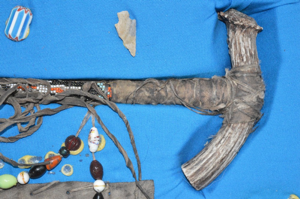 A framed display case lined with blue cloth containing Native American artefacts, including a beaded leather pouch, trade beads, arrowheads, and a hafted tool. View VII
