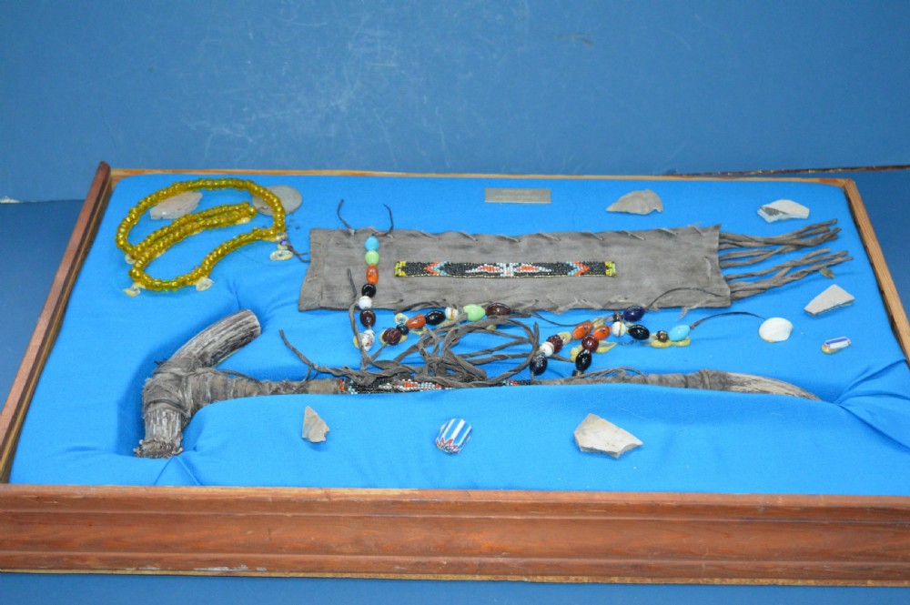 A framed display case lined with blue cloth containing Native American artefacts, including a beaded leather pouch, trade beads, arrowheads, and a hafted tool. View IX