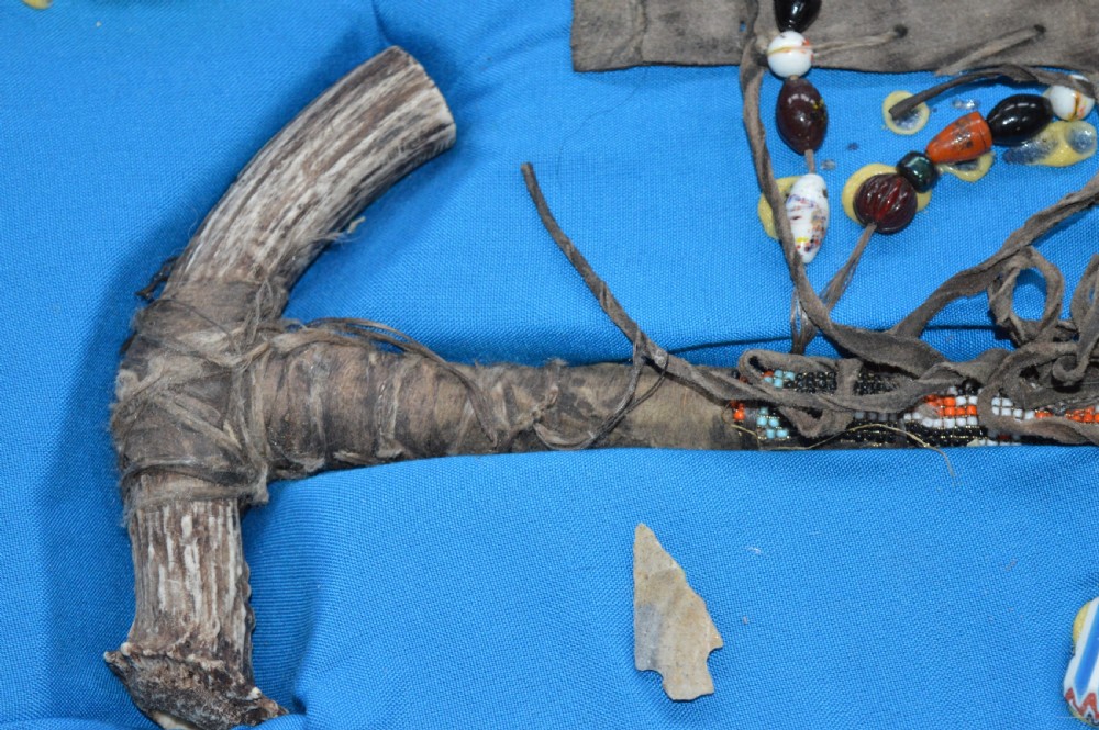 A framed display case lined with blue cloth containing Native American artefacts, including a beaded leather pouch, trade beads, arrowheads, and a hafted tool. View X