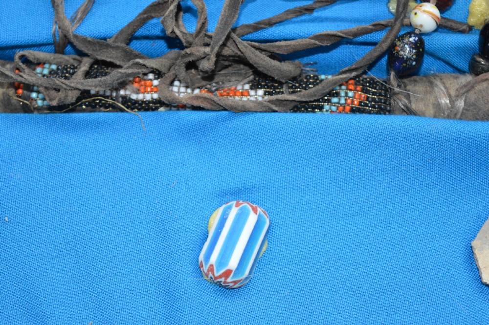 A framed display case lined with blue cloth containing Native American artefacts, including a beaded leather pouch, trade beads, arrowheads, and a hafted tool. View XI