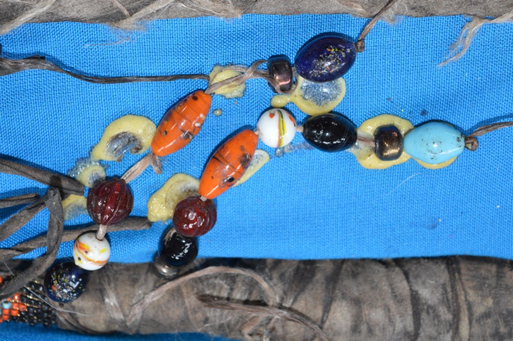 A framed display case lined with blue cloth containing Native American artefacts, including a beaded leather pouch, trade beads, arrowheads, and a hafted tool. View XII