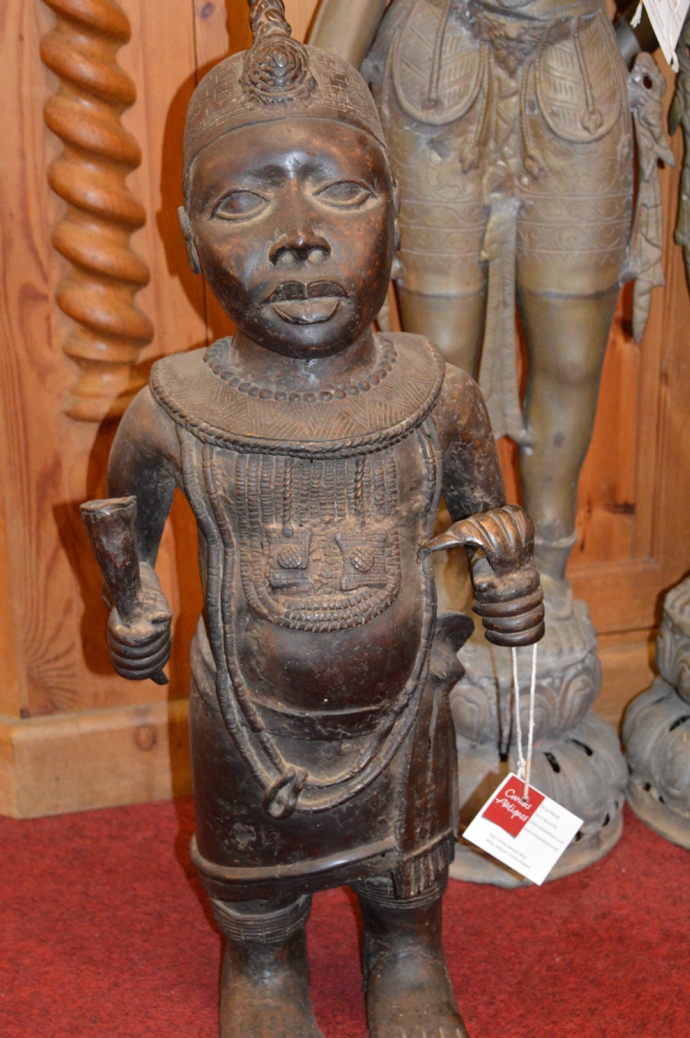 A standing cast bronze West African warrior figure with ceremonial headwear, detailed chest ornamentation, and traditional waist garment.